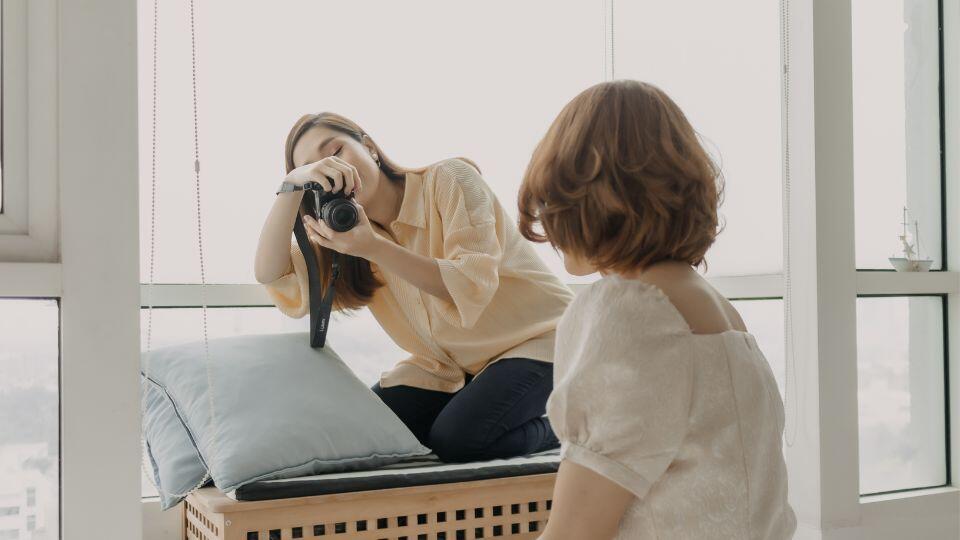 A woman sitting on a bench by a large window takes a photo of another woman sitting nearby. The scene is indoors, with soft natural light and relaxed, casual poses.