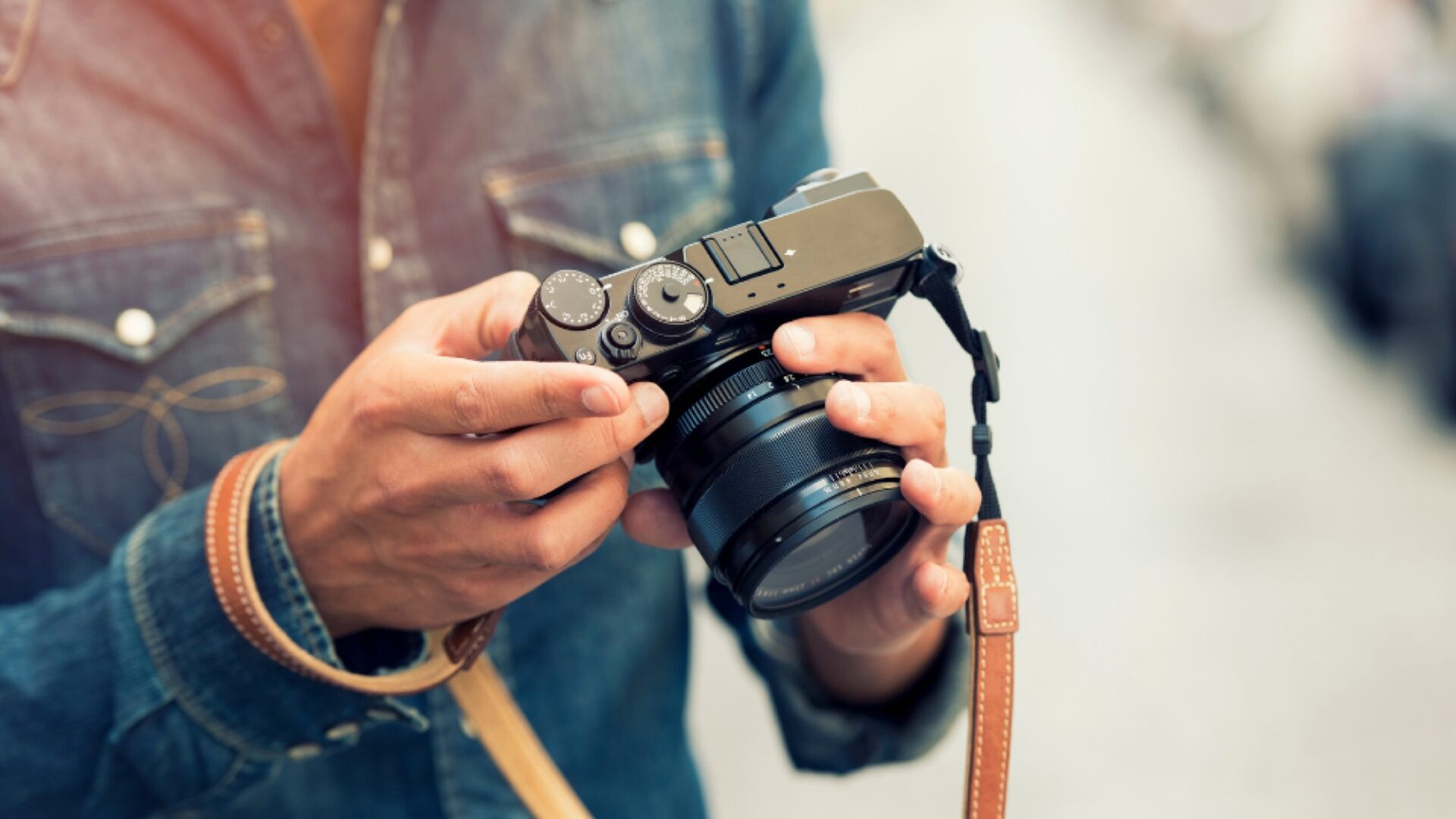 A person wearing a denim shirt holds a black digital camera with both hands, adjusting its settings. The camera has a leather strap attached, and the background is softly blurred.