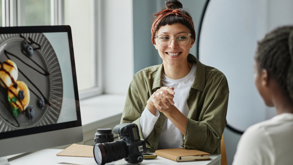 A smiling woman wearing glasses and a headband sits at a desk with a camera and lenses, talking to another person. A computer monitor displays a close-up image of a dessert with sauce and berries.