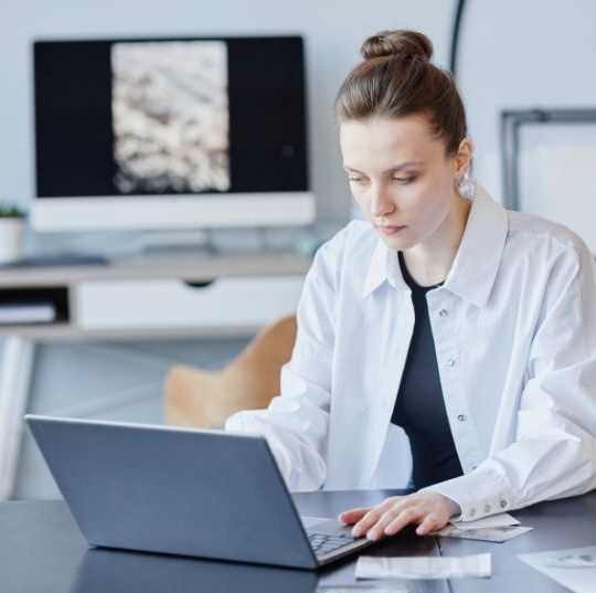 A woman in a white shirt sits at a desk working on a laptop. An office chair and a computer monitor are visible in the background. She appears focused and is using her right hand to type.