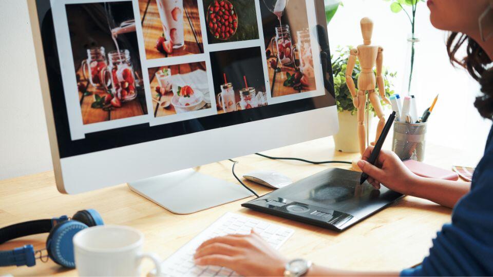 A person using a digital drawing tablet and stylus at a desk with a computer displaying photos of food and drinks. The workspace includes a keyboard, headphones, and a small wooden mannequin.