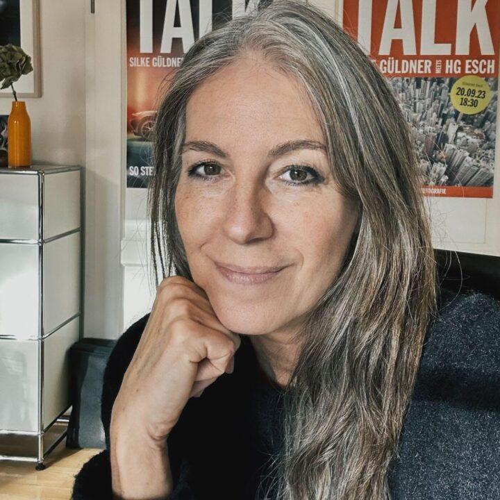 A woman with long gray hair smiles softly at the camera, resting her chin on her hand. She is indoors, with posters and a metal cabinet visible in the background.