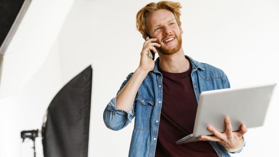 A smiling man with red hair and a beard holds a laptop in one hand and talks on a smartphone. He is wearing a denim shirt over a dark t-shirt, standing in a bright studio with photo equipment in the background.