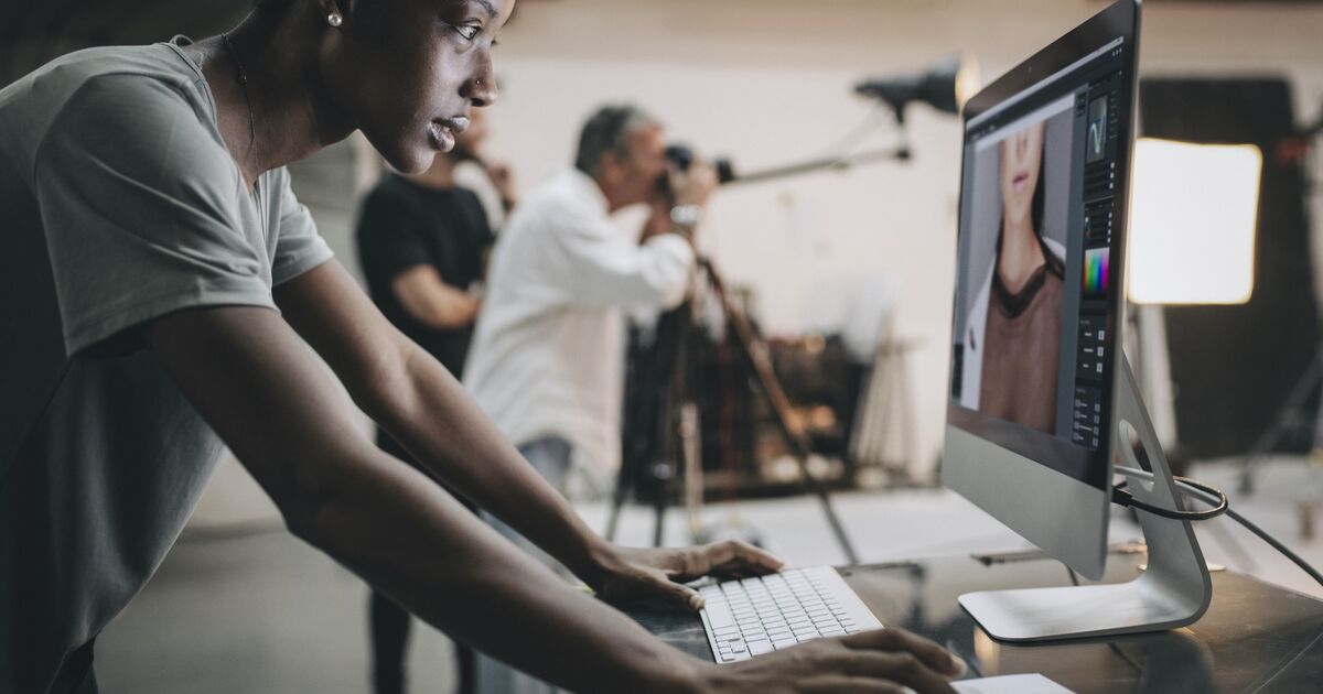A person works on a desktop computer editing a portrait photo, while photographers with cameras and lighting equipment work in the blurred background of a professional studio.