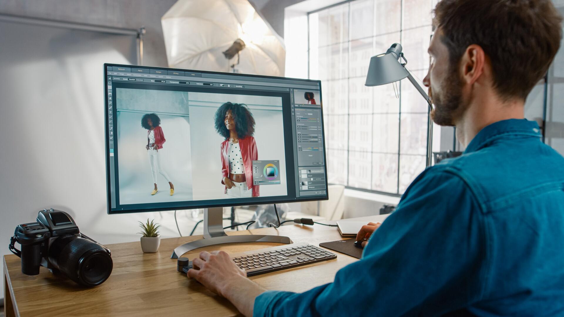 A man at a desk edits fashion photos of a woman on a computer screen. He uses a stylus and graphics tablet, with a camera, keyboard, and potted plant nearby in a well-lit studio.