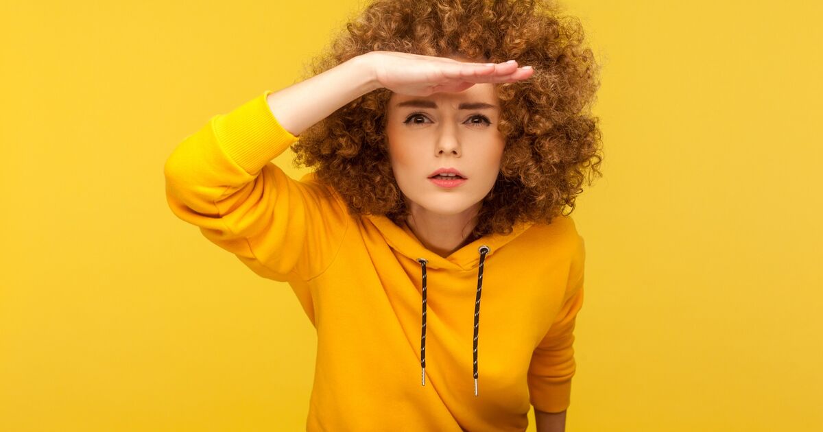 A young person with curly hair wearing a yellow hoodie stands against a yellow background, squinting and shielding their eyes with one hand as if looking into the distance.