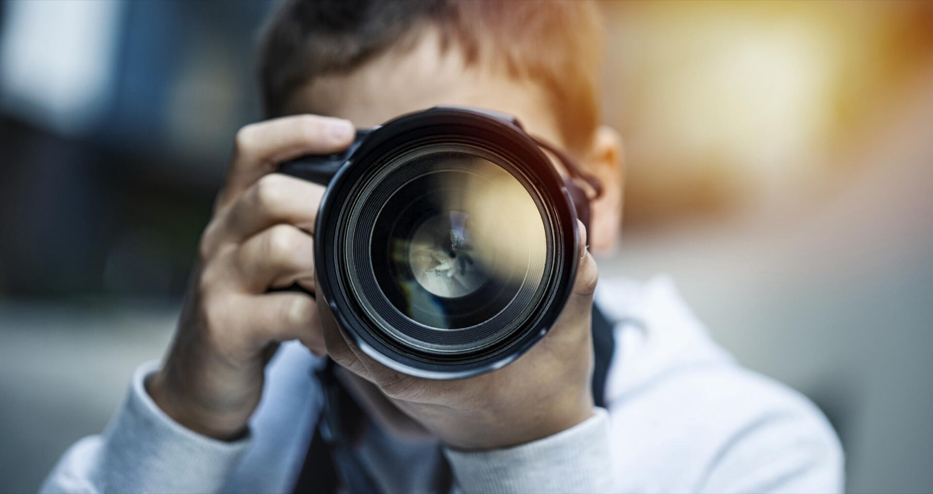A child holds a large camera close to their face, looking through the lens as if taking a photo. The background is softly blurred, and sunlight creates a warm glow in the image.
