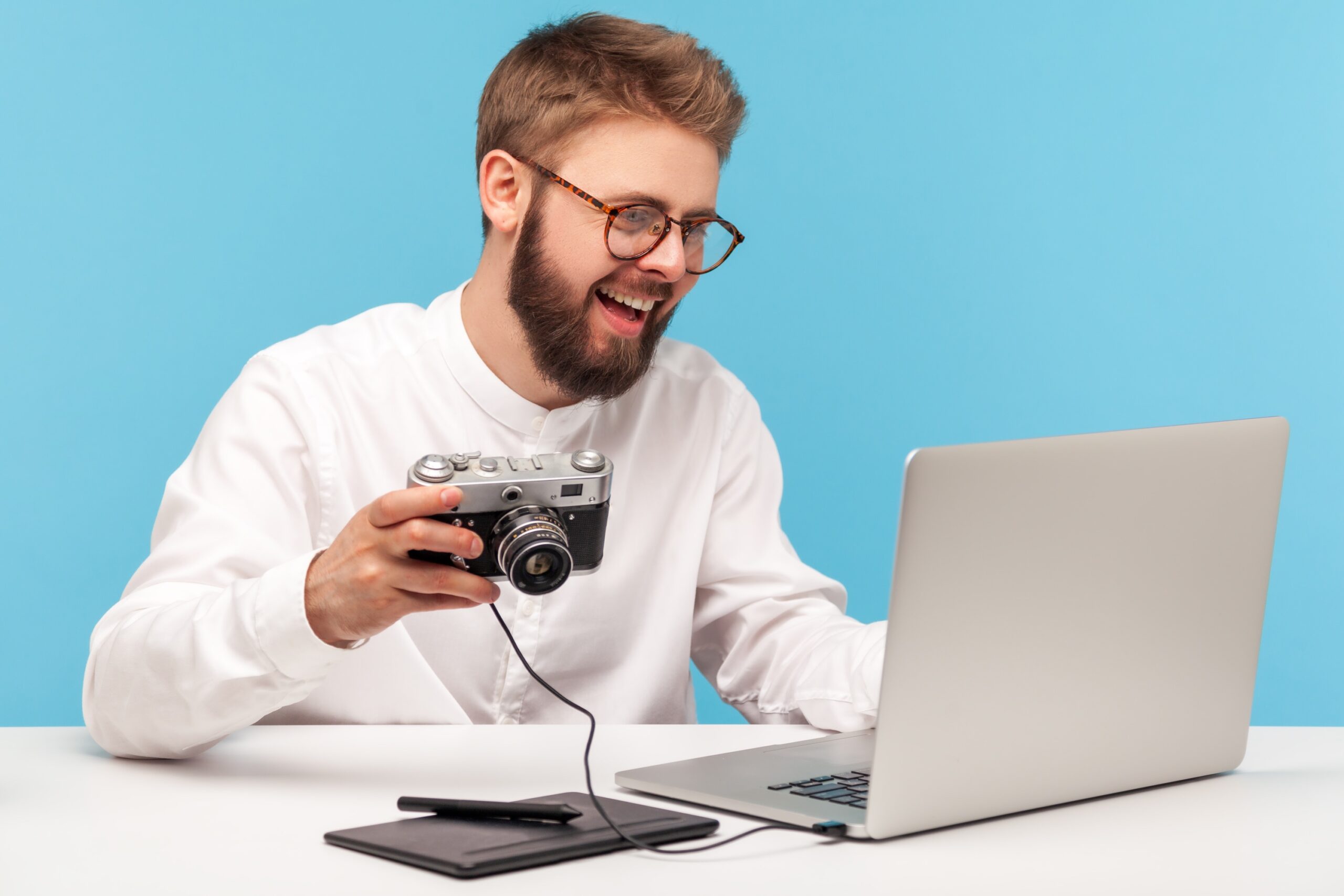 Ein bärtiger Mann mit Brille und weißem Hemd lächelt, während er eine an einen Laptop angeschlossene Kamera auf einem Schreibtisch vor blauem Hintergrund hält.