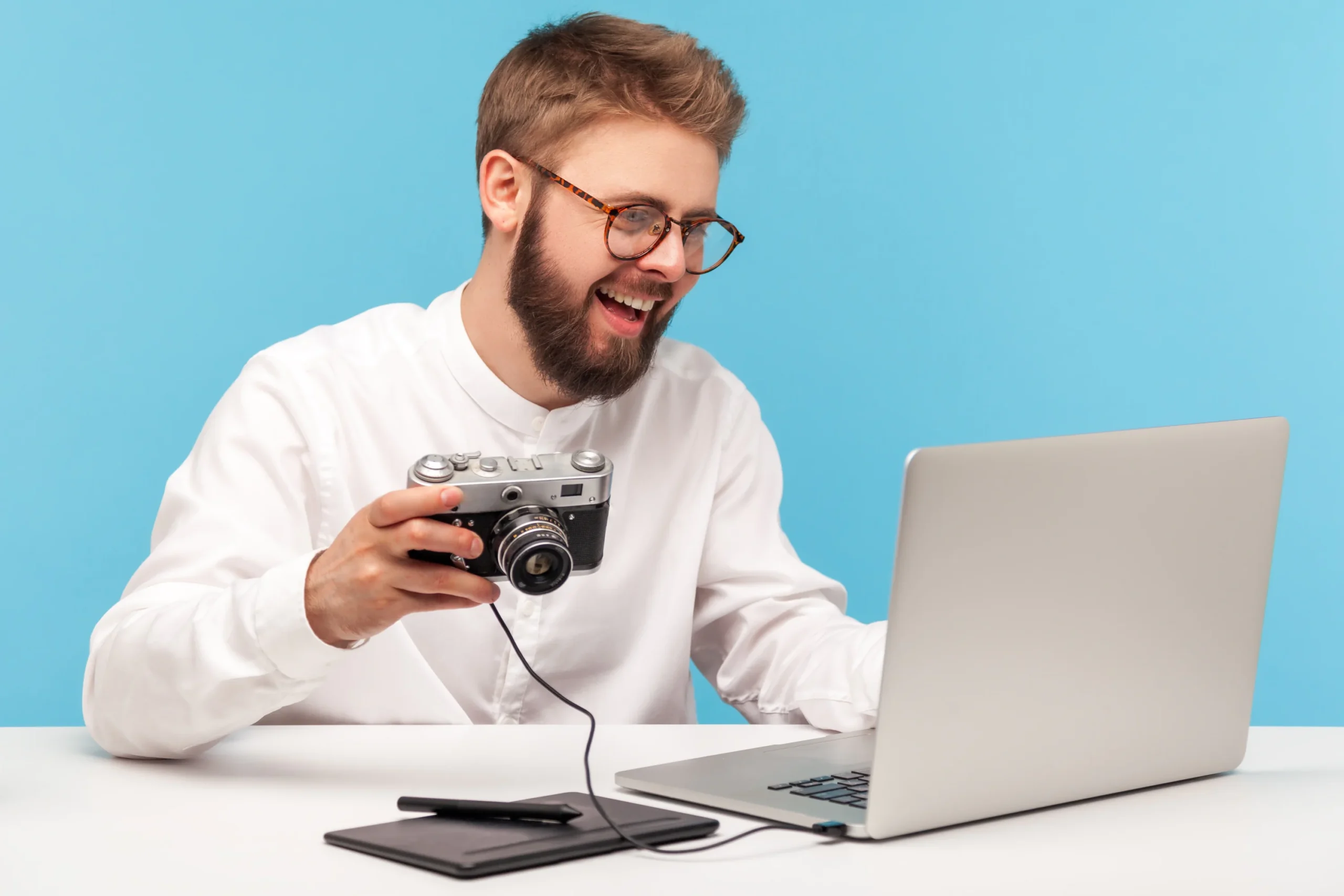 A smiling man with glasses and a beard sits at a desk, holding a camera connected to a laptop. He wears a white shirt and is looking at the screen. A tablet and pen are also on the desk against a blue background.