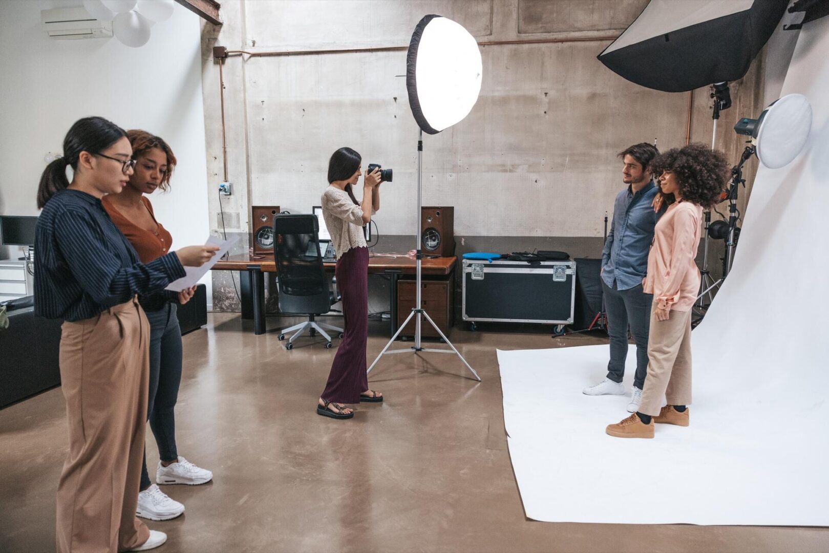 A group of people work in a photo studio; two people pose in front of a white backdrop while a photographer takes their picture and two others look at papers nearby. Studio lights and equipment are visible.