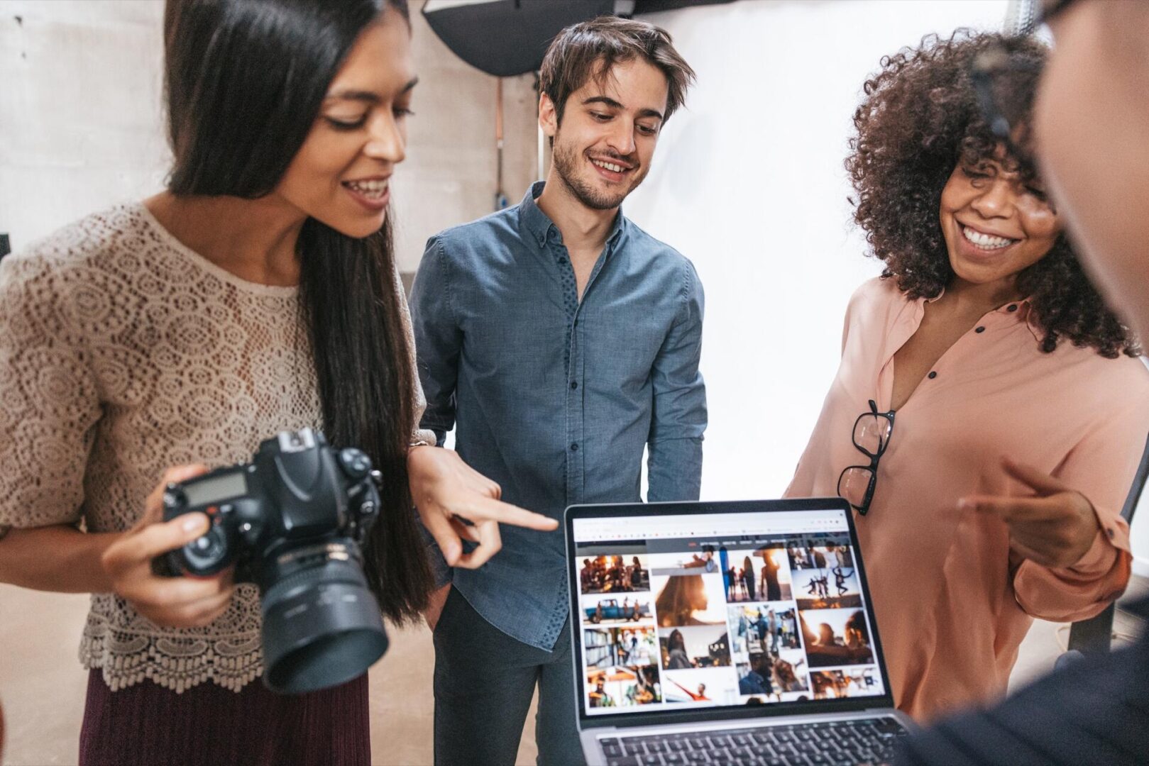 Three people stand together, smiling and pointing at photos displayed on a laptop screen. One person holds a camera, suggesting they are reviewing recent pictures in a bright indoor studio setting.