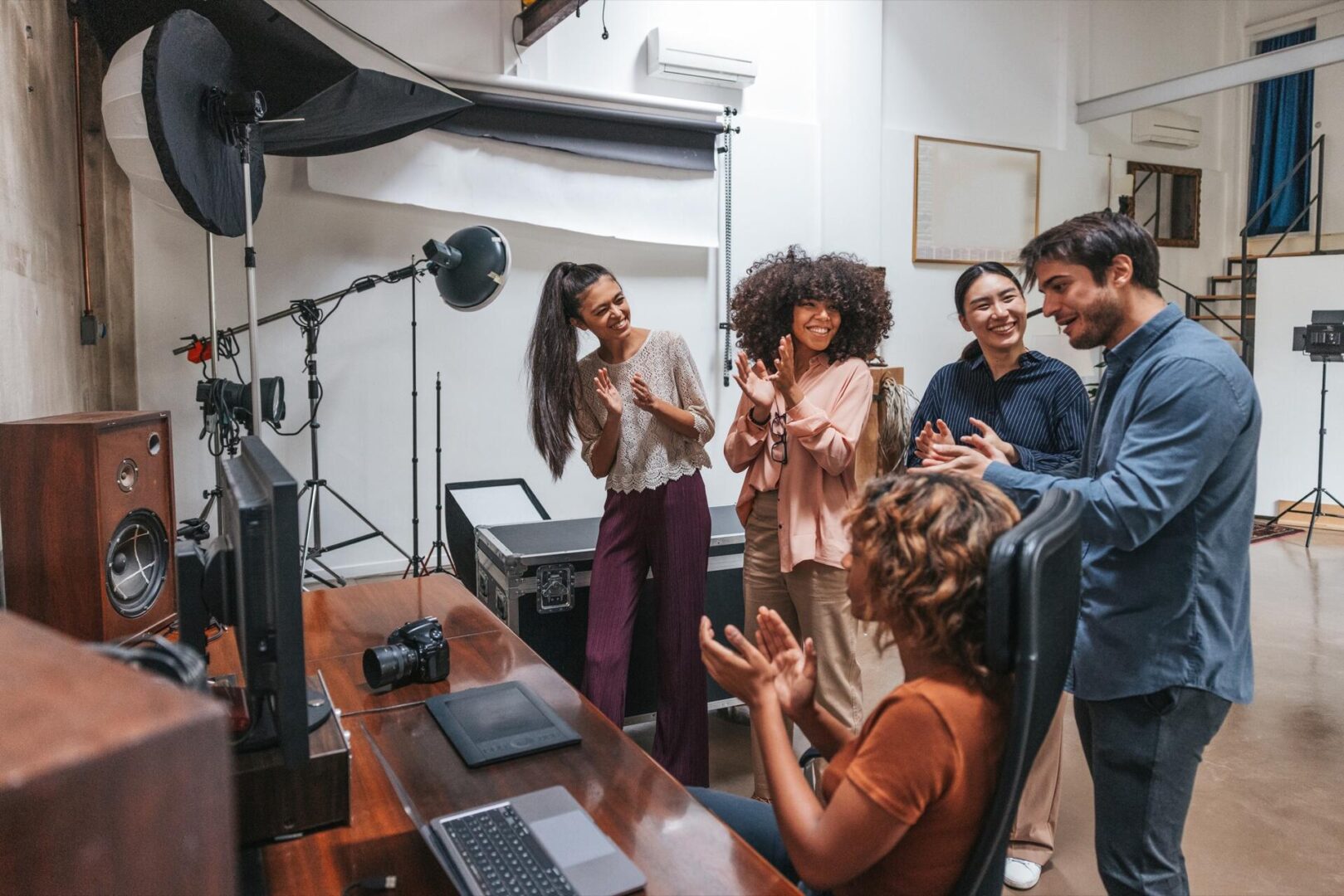 A group of five people in a photography studio are smiling and clapping around a woman seated at a computer desk, celebrating and enjoying a positive moment together.