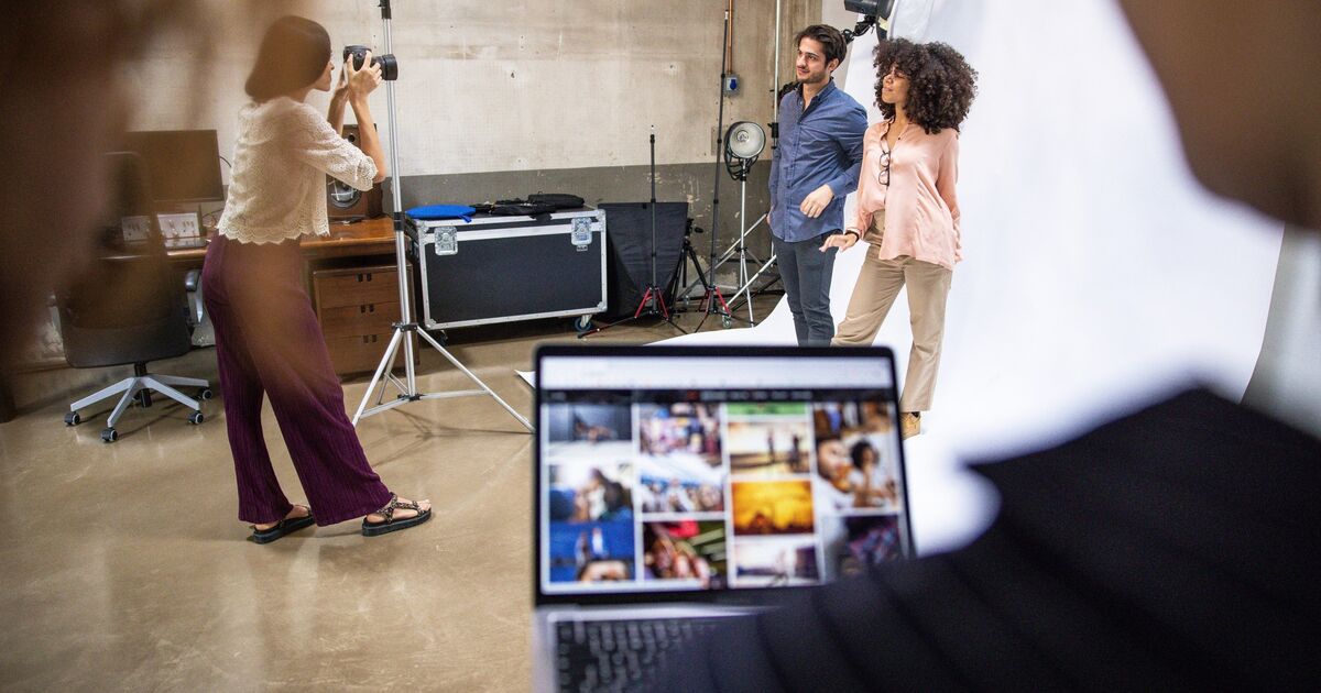 A photographer takes pictures of a smiling man and woman posing together in a studio, while a person in the foreground views photos on a laptop. Studio lights and equipment are visible in the background.
