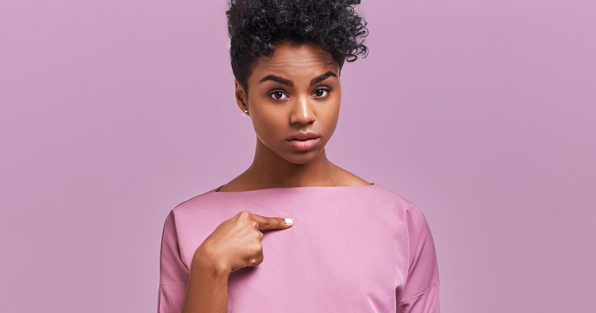 A woman with short curly hair wearing a pink top stands against a matching pink background, looking directly at the camera while pointing to herself with her right index finger.