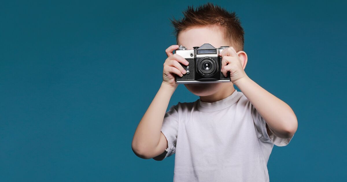 A young child with short brown hair wearing a white t-shirt holds a vintage camera up to their face, standing against a plain blue background.