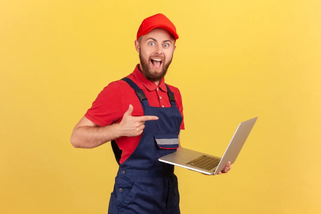 Ein bärtiger Mann mit roter Mütze und Overall zeigt aufgeregt auf einen Laptop, den er in einer Hand hält und vor einem einfarbig gelben Hintergrund steht.