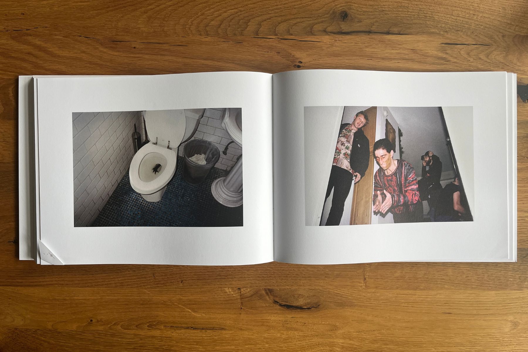 Open photobook on a wooden table, with a photograph of an object in a toilet on the left page and several people gathered in a hallway on the right.