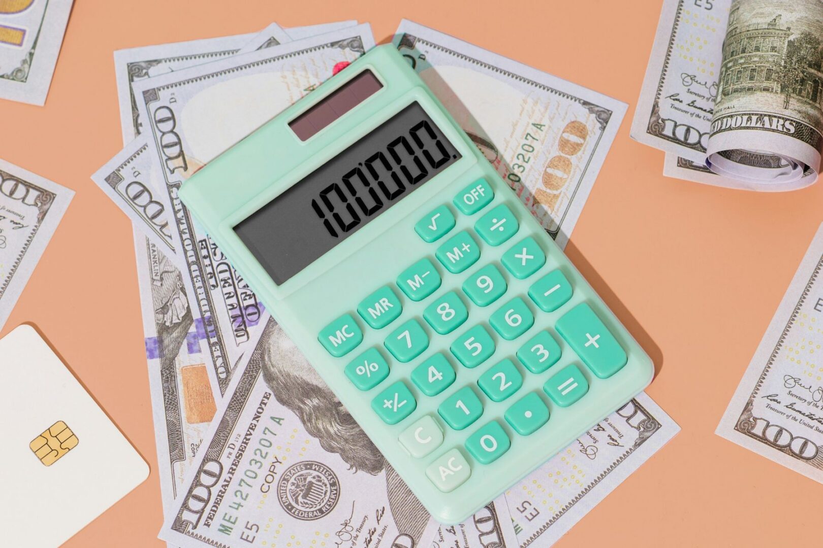 Mint-green calculator showing 100,000, placed on US dollar bills with a credit card on a peach-colored background.