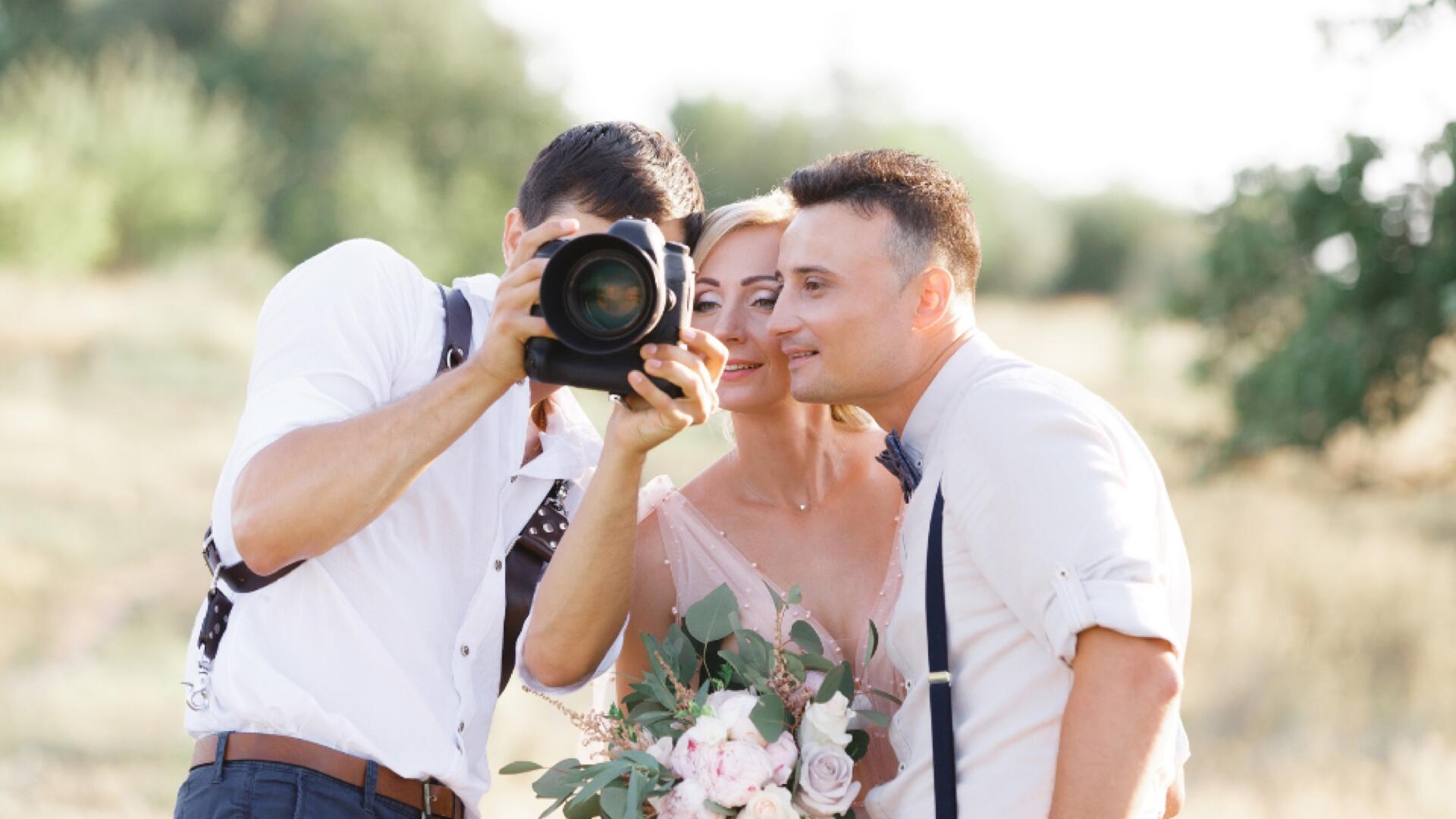 A wedding photographer shows a photo on his camera to a smiling bride and groom holding a bouquet, outdoors in a sunny, green setting.