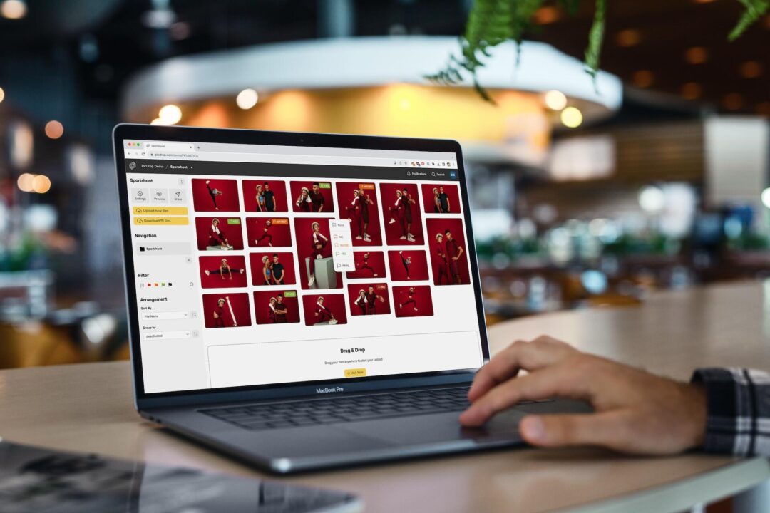 Eine Person nutzt einen Laptop, auf dem eine Fotoverwaltungsanwendung mit mehreren Porträts vor rotem Hintergrund in einem Raster angezeigt wird. Die Hand des Benutzers liegt auf dem Trackpad in einem modernen, hellen Café-Ambiente.