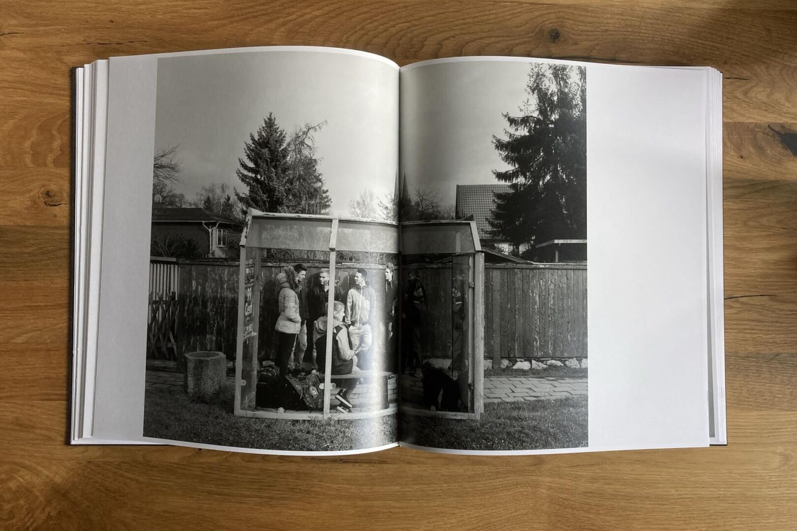 A black and white photo in an open book shows several people gathered inside a small outdoor bus shelter. Trees, a wooden fence, and houses are visible in the background. The book rests on a wooden table.