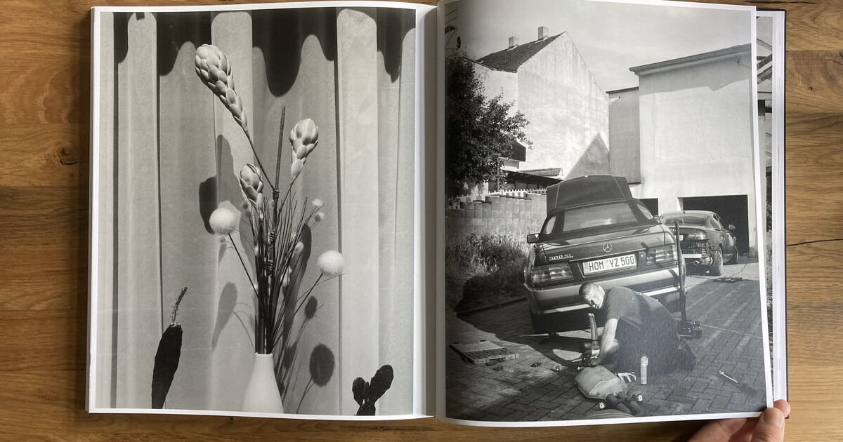 A book is open to two black-and-white photos: the left page shows a vase with flowers and shadows on a striped wall; the right page shows a person kneeling by a car parked in a driveway in front of white buildings.