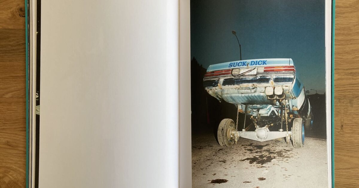 A photo book open to a page showing the rear of a battered car with large tires; the cars back has SUCK DICK written on it in large letters. The background is dark and the ground appears dirty.