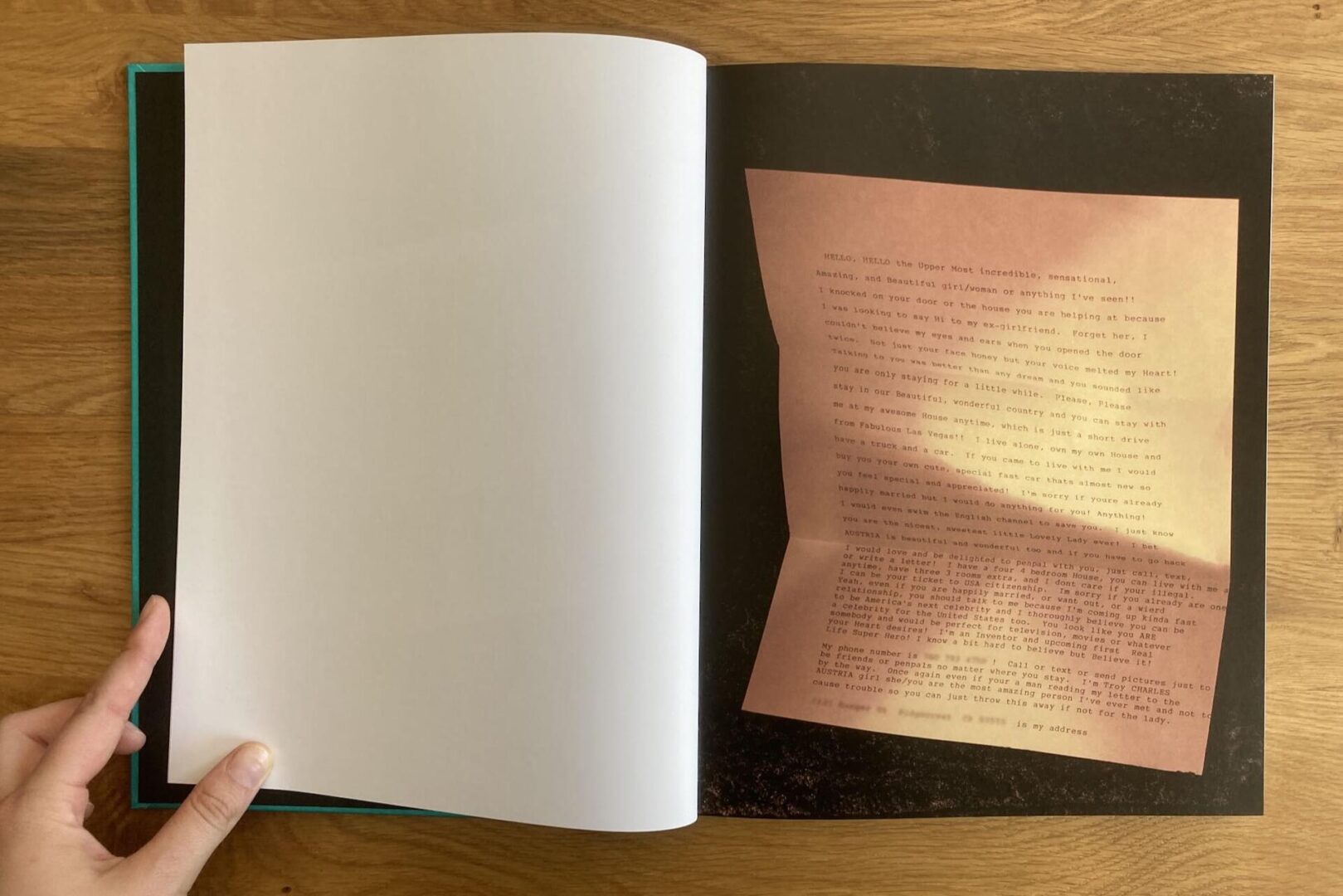A hand holds open a book to a page with a color photo of a pink typed letter on the right side, placed on a dark textured background. The left page is blank. The book rests on a wooden surface.