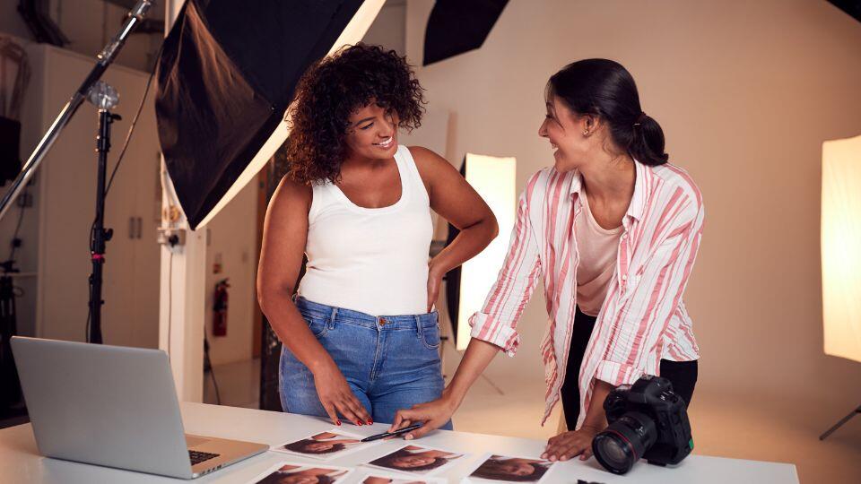 Two women smile and discuss printed photos on a table in a photography studio. One holds a camera and the other is pointing at images. Studio lights, a laptop, and photo equipment are visible in the background.