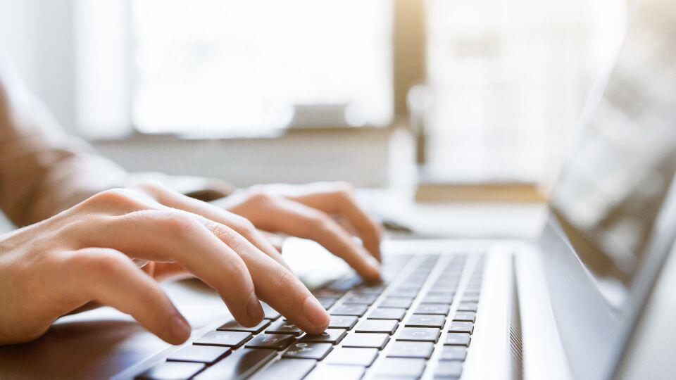 Close-up of hands typing on a laptop keyboard in a bright room with sunlight streaming through a window in the background.
