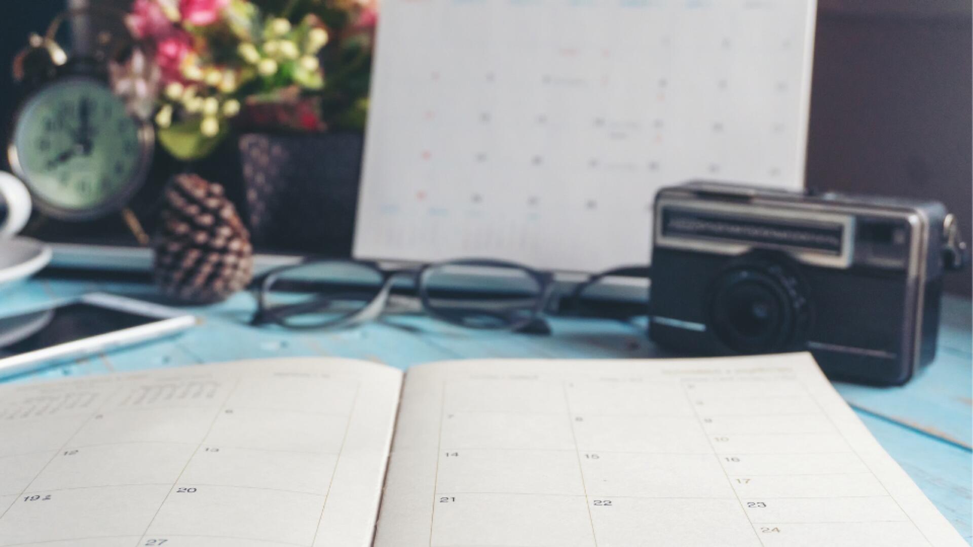 An open planner sits on a blue desk alongside a pair of glasses, a vintage camera, a clock, a smartphone, and a blurred calendar in the background, with plants adding a decorative touch.