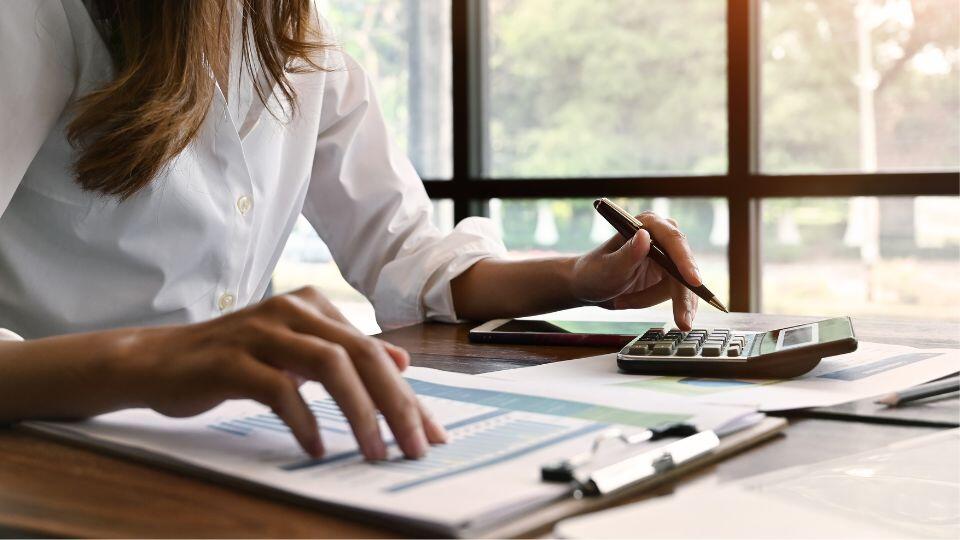 A person in a white shirt sits at a desk using a calculator and reviewing financial documents, with sunlight coming through large windows in the background.