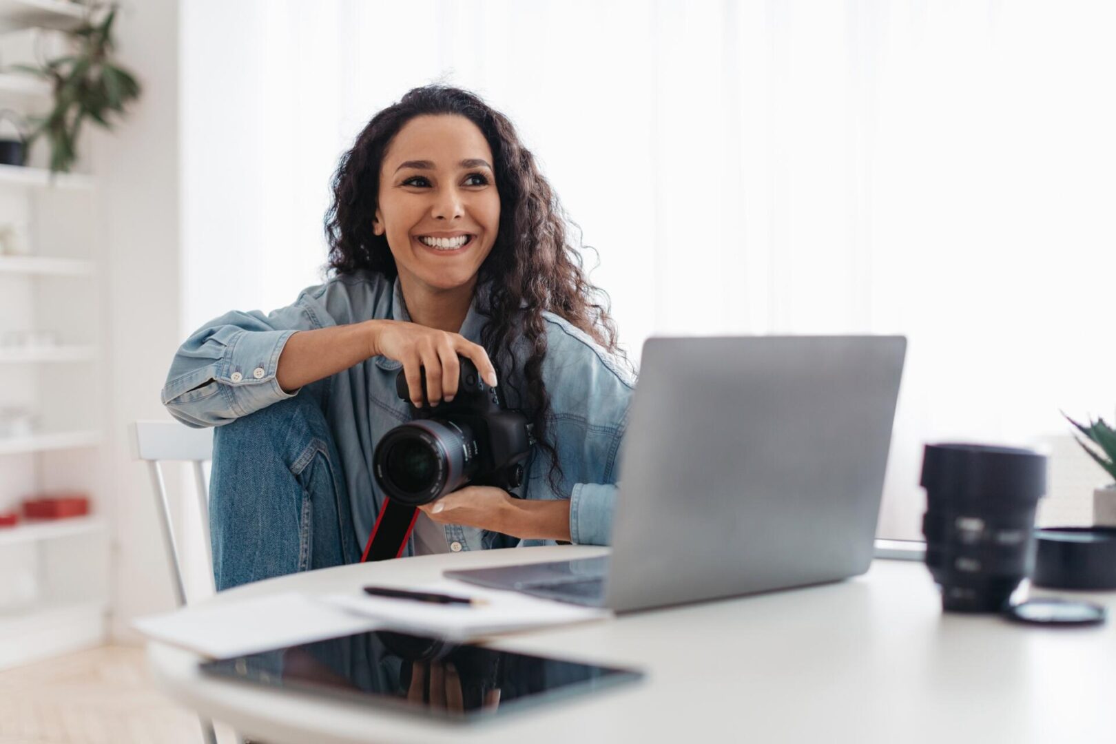 A smiling woman with curly hair sits at a desk holding a camera, with a laptop, tablet, camera lens, and papers in front of her. Bright, natural light fills the room.
