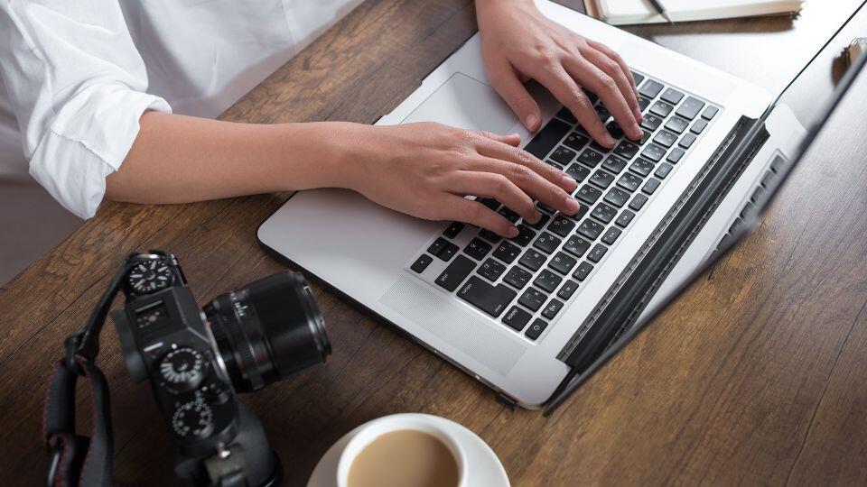 A person types on a laptop at a wooden desk with a camera, a notebook, and a cup of coffee nearby.
