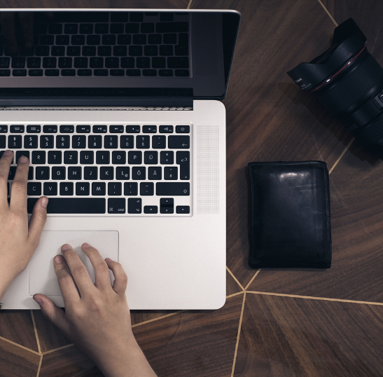 A person’s hands typing on a laptop keyboard, with a camera and a closed black wallet placed on a wooden table nearby.