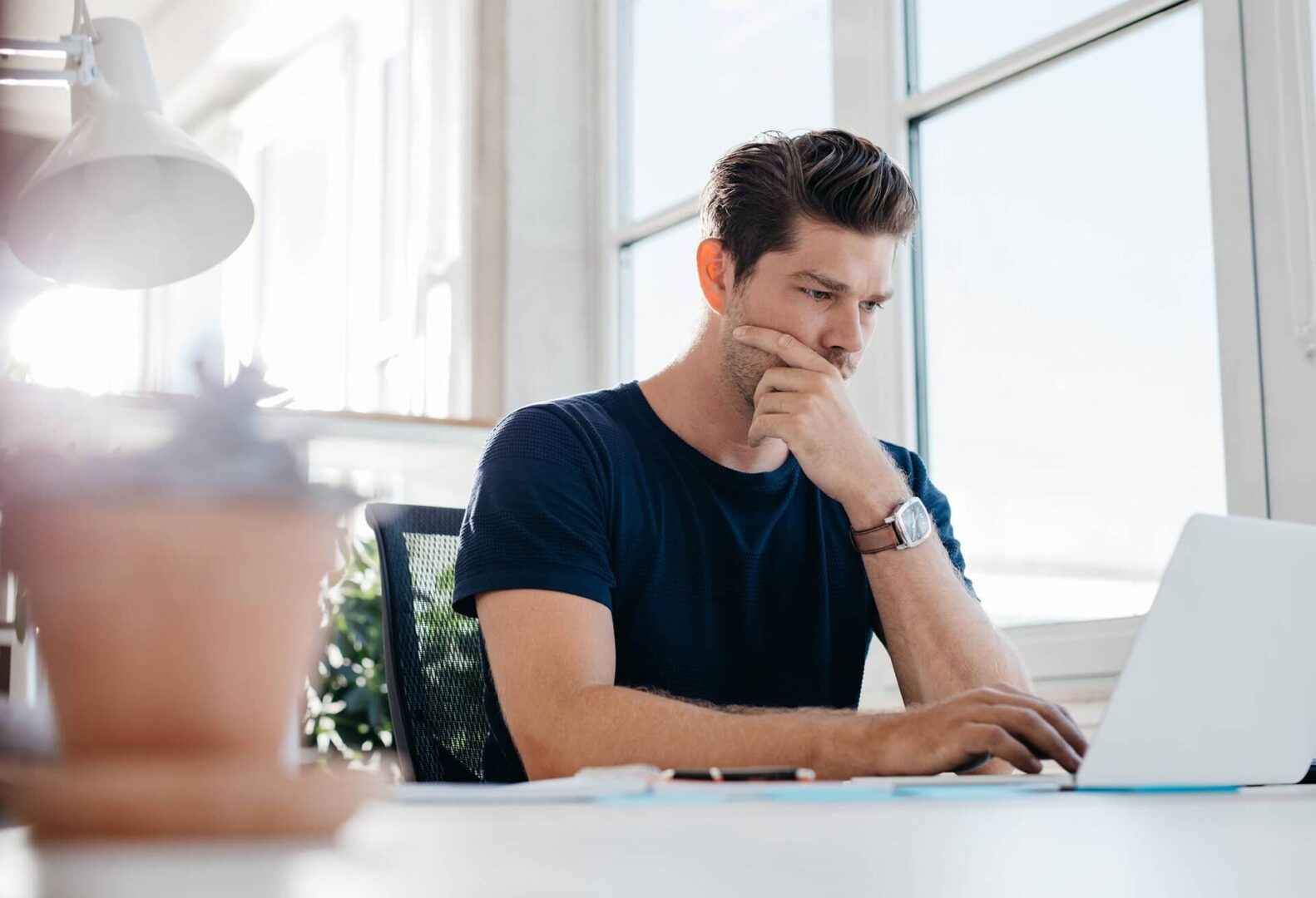 A man sits at a desk in a bright office, looking thoughtfully at a laptop. He rests his chin on his hand, appearing focused and deep in concentration. A plant and lamp are visible in the foreground.