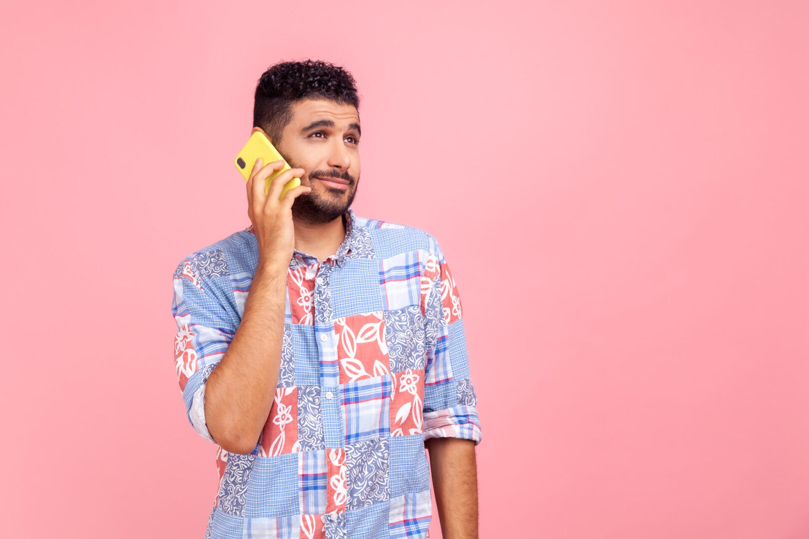 A man with short curly hair and a trimmed beard holds a yellow smartphone to his ear, looking uncertain or confused, standing against a plain pink background.