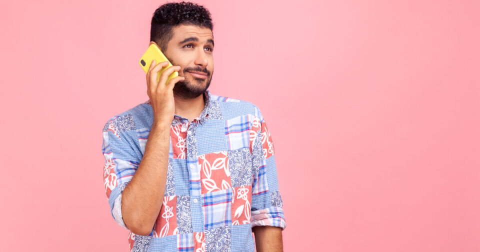 A man with short curly hair and a beard holds a yellow phone to his ear, looking uncertain or skeptical. He wears a colorful patterned shirt and stands against a plain pink background.