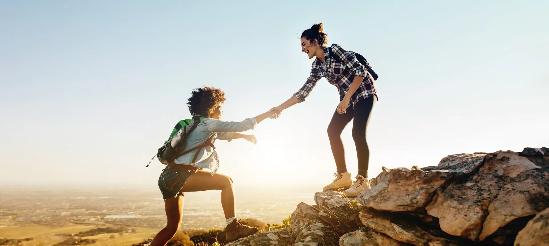 A person helps another climb up a rocky hill at sunrise or sunset, both smiling and dressed casually with backpacks, with a scenic view of a city and sky in the background.