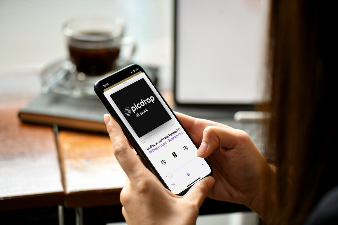 Eine Person hält ein Smartphone, auf dem eine Podcast-Folge mit dem Titel „Picdrop“ läuft, bei der Arbeit. Im Hintergrund stehen eine verschwommene Tasse Kaffee und ein Laptop auf dem Holztisch.