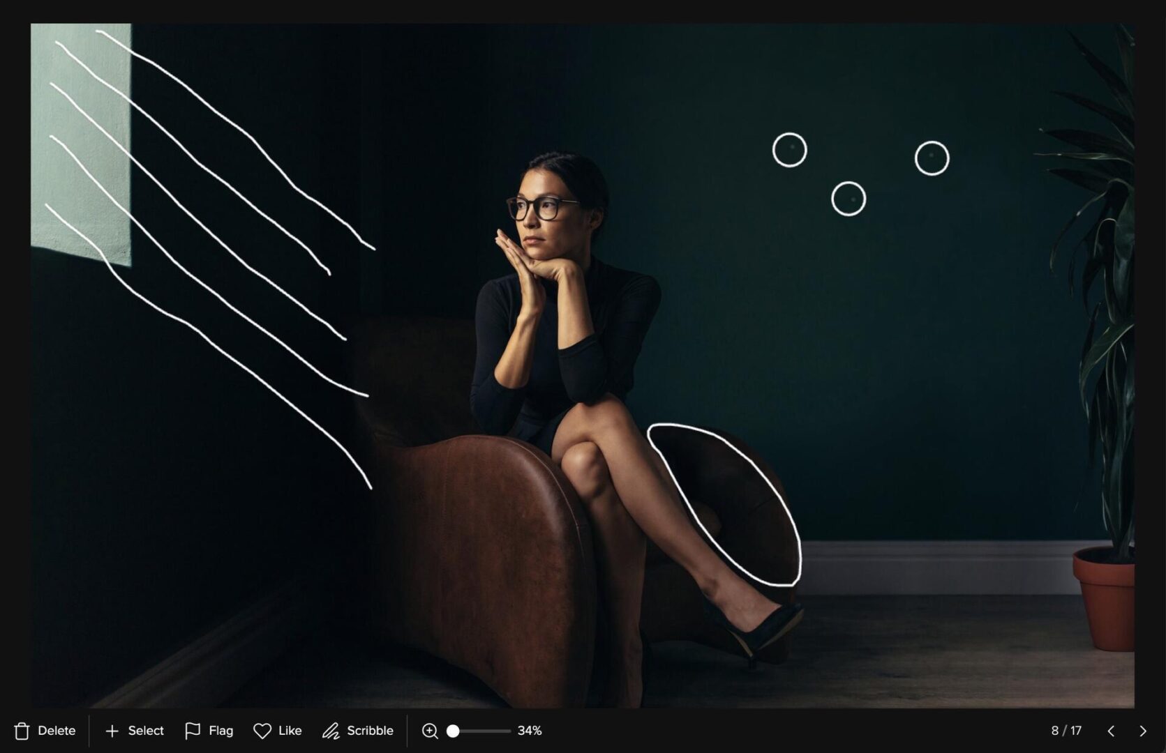 A woman in glasses and a black dress sits thoughtfully on a brown chair in a dimly lit room. White doodles, including lines and circles, are drawn over the image, adding an artistic touch.