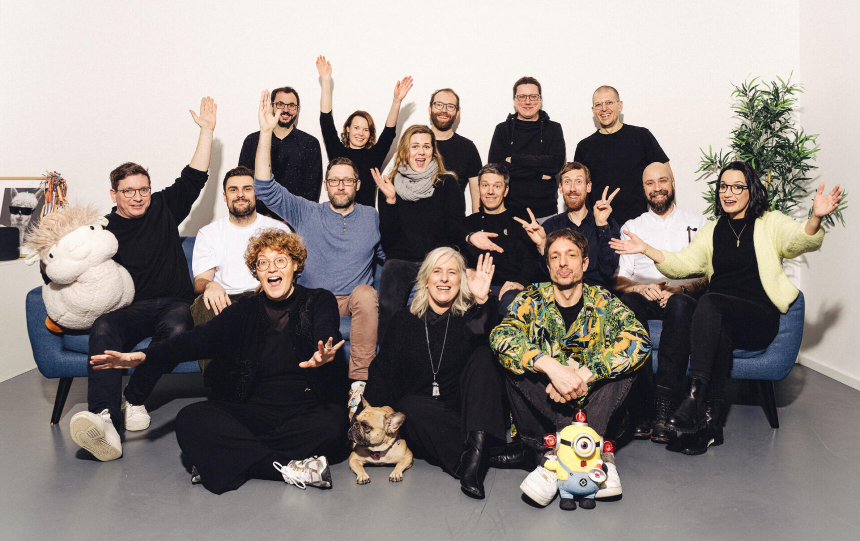 A group of 17 people pose cheerfully indoors, some sitting and some standing by a blue couch. There’s a plush sheep, a small dog, and Minion and Pokémon plush toys in front, with a plant on the right.