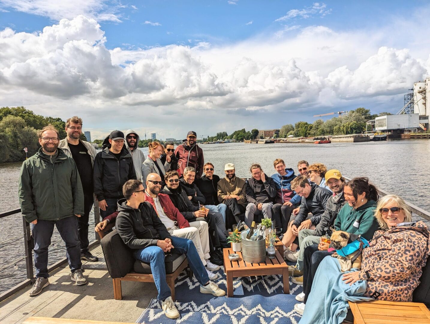 Eine Gruppe von etwa 20 Personen und ein kleiner Hund sitzen und stehen gemeinsam auf einem Bootsdeck und lächeln in die Kamera. Im Hintergrund sind ein Fluss, Grünflächen, Gebäude und ein teilweise bewölkter Himmel zu sehen.