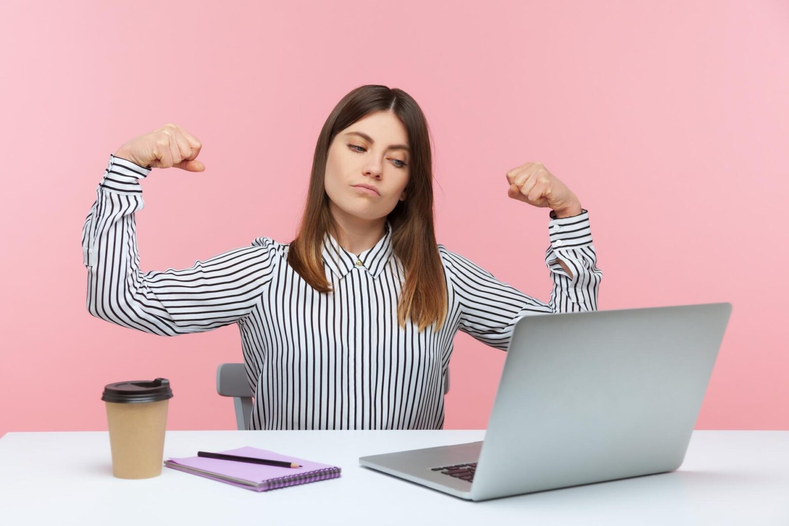 A woman in a striped shirt sits at a desk with a laptop, notebook, pen, and coffee cup, flexing her arms confidently and smiling slightly against a pink background.