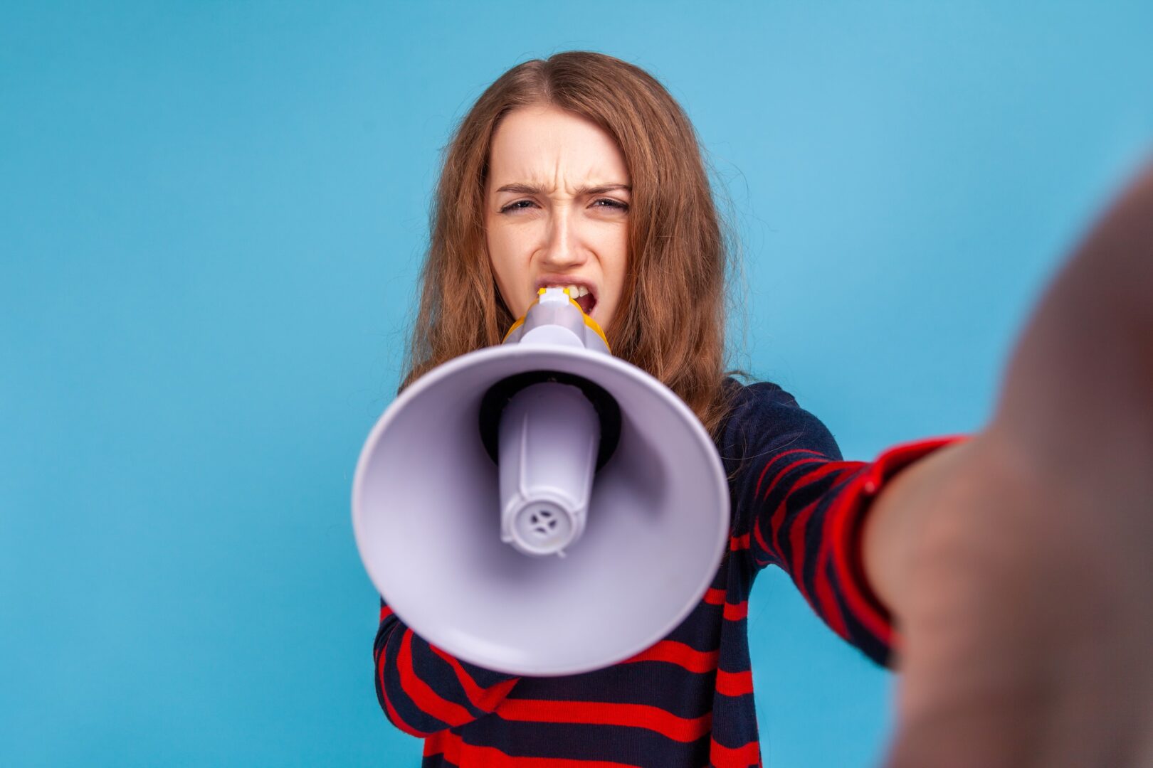 A young woman with long brown hair, wearing a red and navy striped shirt, shouts into a megaphone while holding it toward the camera against a blue background. Her expression appears intense or frustrated.