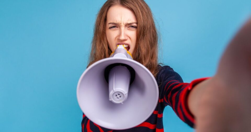 Young woman with long brown hair, wearing a dark top with red stripes, holds a megaphone towards the camera and appears to shout, with a serious expression, against a solid blue background.