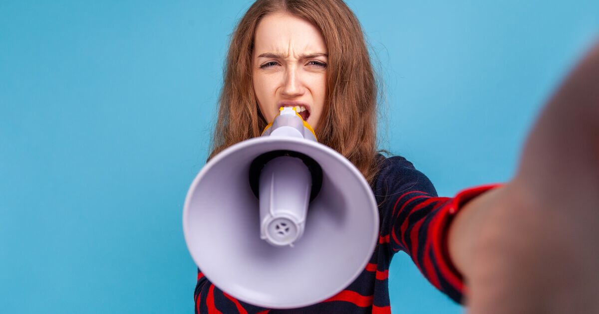 Young woman with long brown hair, wearing a dark top with red stripes, holds a megaphone towards the camera and appears to shout, with a serious expression, against a solid blue background.