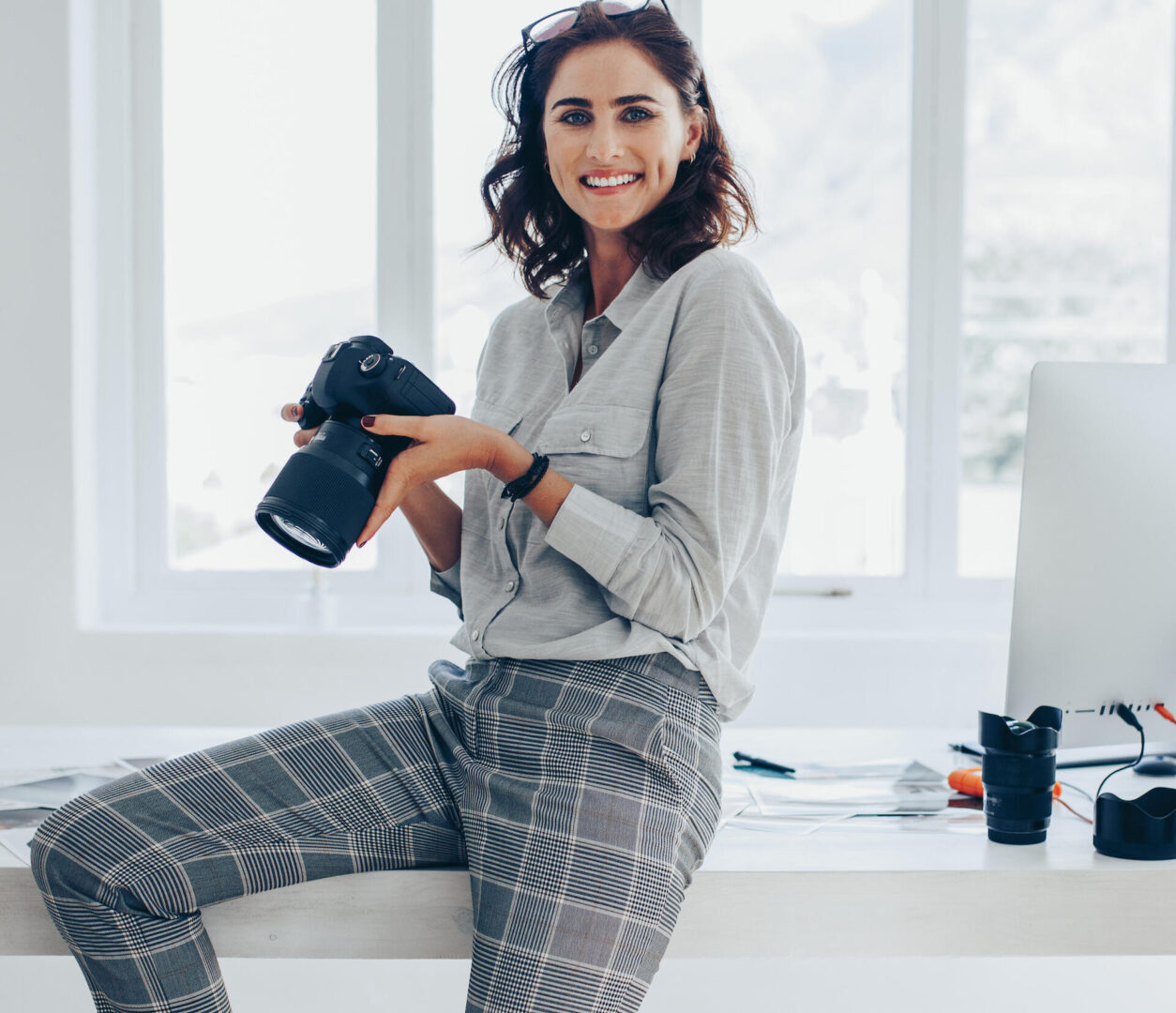 A woman in a gray shirt and plaid pants sits on a desk, smiling and holding a camera. Behind her are large windows and an office setup with a computer, lens, and stationery on the desk.