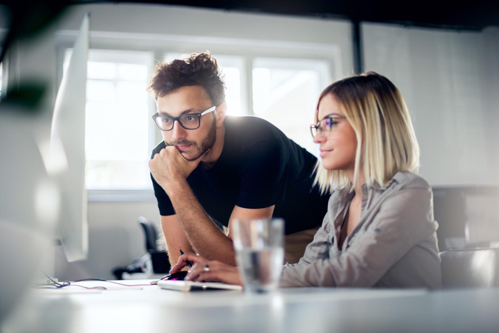 Two people wearing glasses work together at a computer in a bright office. One person sits and types while the other stands beside them, looking at the screen intently. A glass of water is on the desk.