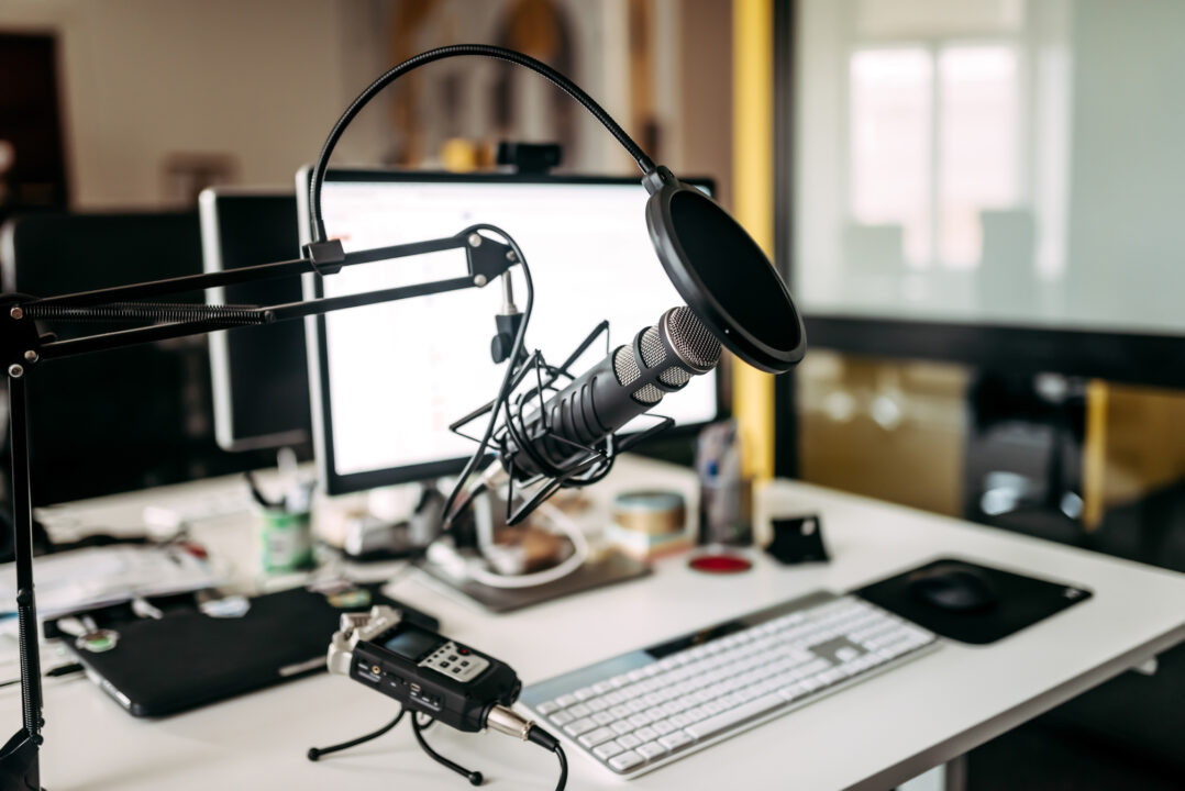 Ein Tischmikrofon mit Pop-Filter ist auf einem Galgenarm vor einem Computermonitor auf einem modernen Büroschreibtisch aufgestellt, umgeben von Aufnahmegeräten, einer Tastatur und anderen Büromaterialien.